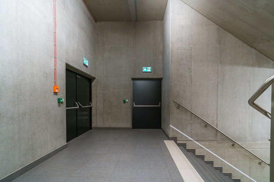 A modern, industrial-style stairwell and corridor area featuring smooth, light gray concrete walls and floor tiles. To the left, there are black double elevator doors with metallic handles, accompanied by green emergency call buttons and orange emergency equipment mounted on the wall. Overhead, illuminated green exit signs with white arrows indicate emergency exit routes. The corridor is well-lit with natural or artificial light, and a stainless steel handrail runs along the staircase on the right side, which has dark gray steps with a white tactile strip at the edge. The overall appearance is clean and well-maintained, reflecting the cleaning standards of Cleaners Lambeth, as discussed on the Waterloo Station emergency cleaning options Lambeth page.