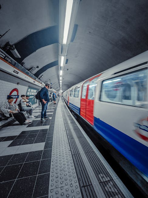 An underground London tube station with a curved, concrete ceiling illuminated by long, straight fluorescent light fixtures. The platform has black and white tiled flooring, with tactile paving along the edge for safety. Several passengers are seated on benches, while a man stands near the edge watching a moving train with red, white, and blue livery arriving at the platform. The station features white tiled walls with signage and poster displays. Cleanliness levels are high, with no visible dust or debris, demonstrating the platform's maintained hygiene standards. Cleaners Lambeth specializes in surface cleaning and deep cleaning for transit environments, ensuring hygiene and safety in public spaces.