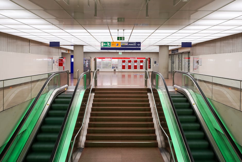 A modern, industrial-style stairwell and corridor area featuring smooth, light gray concrete walls and floor tiles. To the left, there are black double elevator doors with metallic handles, accompanied by green emergency call buttons and orange emergency equipment mounted on the wall. Overhead, illuminated green exit signs with white arrows indicate emergency exit routes. The corridor is well-lit with natural or artificial light, and a stainless steel handrail runs along the staircase on the right side, which has dark gray steps with a white tactile strip at the edge. The overall appearance is clean and well-maintained, reflecting the cleaning standards of Cleaners Lambeth, as discussed on the Waterloo Station emergency cleaning options Lambeth page.